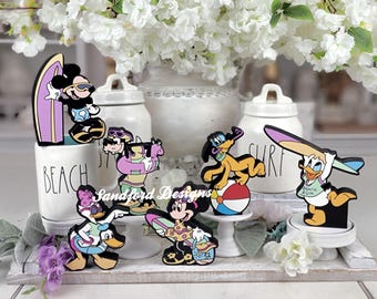 May include: A collection of colorful Disney character figurines, including Mickey Mouse, Minnie Mouse, Donald Duck, and Pluto, each with a surfboard or beach ball. The figurines are set against a backdrop of white ceramic jars and flowers.
