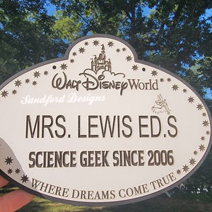 May include: A white oval sign with a black border and black text. The sign has a black and white image of Cinderella's castle and the text 'WALT DISNEY World' at the top. The sign also has the text 'Sandford Designs', 'MRS. LEWIS ED.S', 'SCIENCE GEEK SINCE 2006', and 'WHERE DREAMS COME TRUE'.