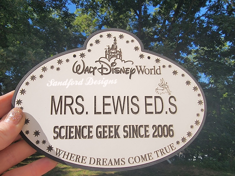 May include: A white oval sign with a black border and black text. The sign has a black and white image of Cinderella's castle and the text 'WALT DISNEY World' at the top. The sign also has the text 'Sandford Designs', 'MRS. LEWIS ED.S', 'SCIENCE GEEK SINCE 2006', and 'WHERE DREAMS COME TRUE'.