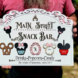May include: White wooden sign with black and pink Mickey Mouse characters and the words 'Main Street Snack Bar' in black. The sign also has the words 'Drinks Popcorn Candy' and 'the origin of happiness, open 24/7' in black.
