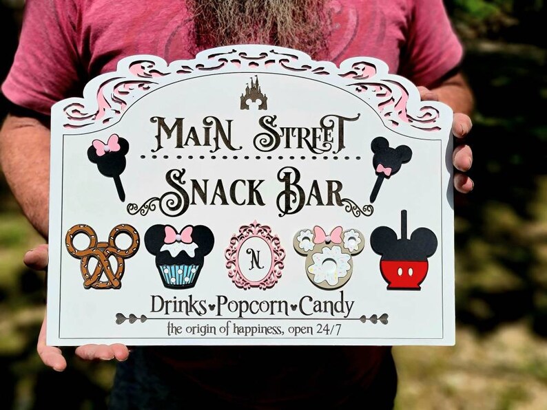 May include: White wooden sign with black and pink Mickey Mouse characters and the words 'Main Street Snack Bar' in black. The sign also has the words 'Drinks Popcorn Candy' and 'the origin of happiness, open 24/7' in black.