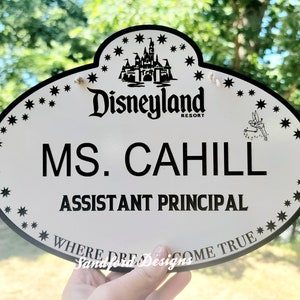 May include: White oval name tag with black text and a black and white image of Sleeping Beauty's castle. The text reads 'Disneyland Resort', 'MS. CAHILL', 'ASSISTANT PRINCIPAL', and 'WHERE DREAMS COME TRUE'. The name tag has a black border with small black stars around the edge.