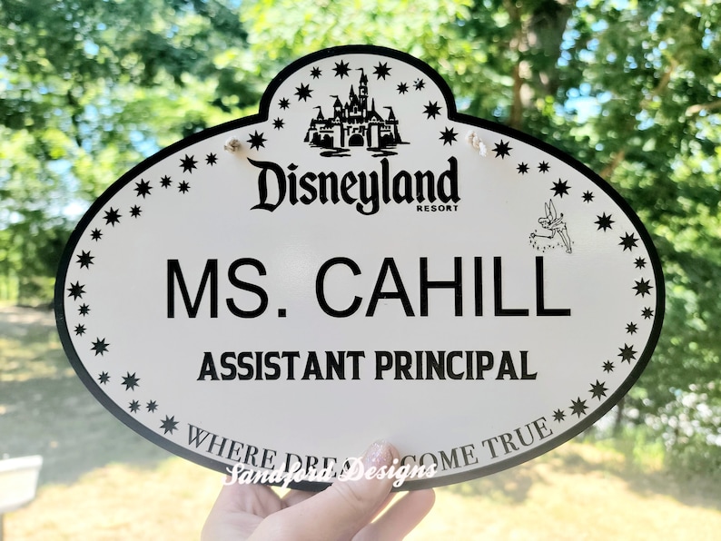 May include: White oval name tag with black text and a black and white image of Sleeping Beauty's castle. The text reads 'Disneyland Resort', 'MS. CAHILL', 'ASSISTANT PRINCIPAL', and 'WHERE DREAMS COME TRUE'. The name tag has a black border with small black stars around the edge.
