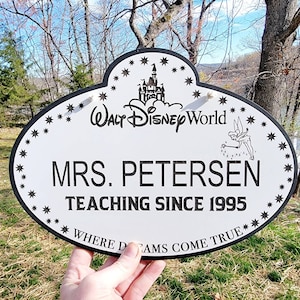 Może przedstawiać: Owalny biały znak z czarną obw&oacute;dką i napisem "MRS. PETERSEN UCZY OD 1995". Zawiera logo Walt Disney World, Dzwoneczka i akcenty gwiazd. Znak głosi "WHERE DREAMS COME TRUE".