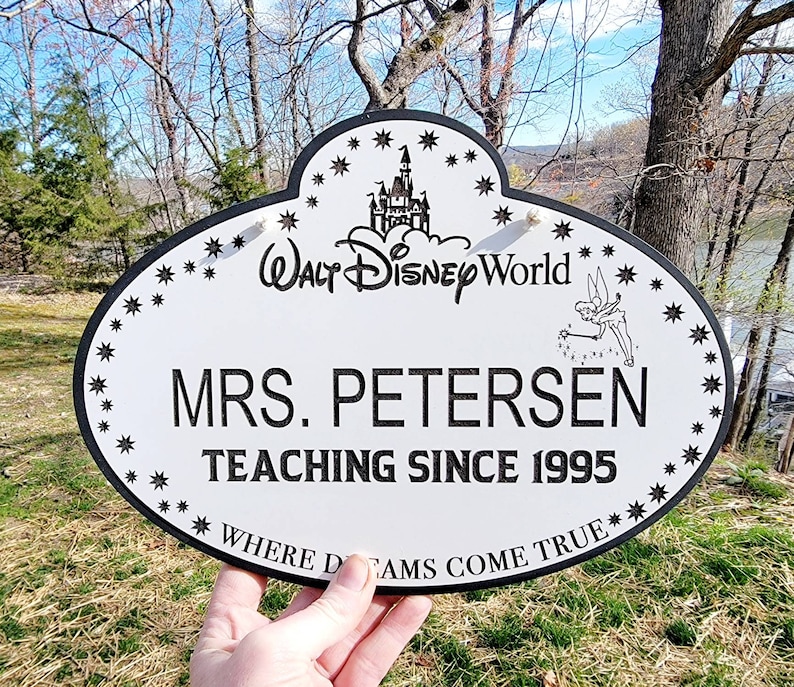 Może przedstawiać: Owalny biały znak z czarną obw&oacute;dką i napisem "MRS. PETERSEN UCZY OD 1995". Zawiera logo Walt Disney World, Dzwoneczka i akcenty gwiazd. Znak głosi "WHERE DREAMS COME TRUE".