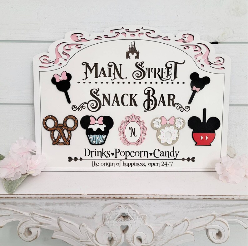 May include: A white wooden sign with pink detailing, reading "Main Street Snack Bar." The sign features Mickey Mouse and Minnie Mouse-themed food icons, such as a pretzel, cupcake, and ice cream. The text "Drinks, Popcorn, Candy" is also included.