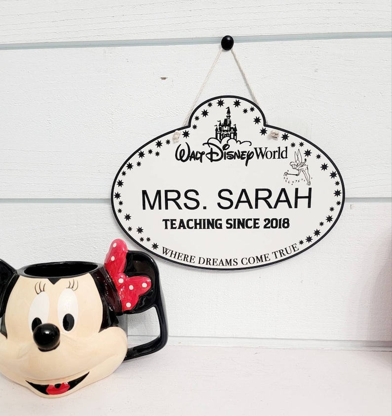 Może przedstawiać: Biały owalny znak z czarnym obramowaniem i napisem "MRS. SARAH TEACHING SINCE 2018" oraz logo "WALT DISNEY WORLD". Kubek Myszki Minnie z czerwoną kokardą znajduje się na pierwszym planie. Znak wisi na białej ścianie.