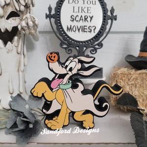 May include: A Halloween-themed decoration featuring a cartoon dog wearing a ghost costume and a pumpkin nose. The dog is in front of a sign that says "Do You Like Scary Movies?" and a witch hat on a hay bale.