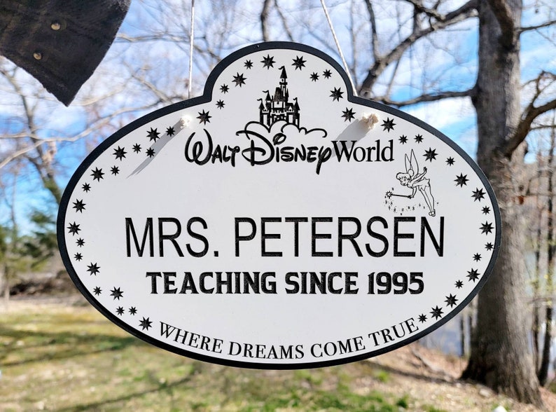 May include: A white oval name tag with a black border. The tag has a black and white image of Cinderella's castle and the text 'Walt Disney World' at the top. The tag also has a black and white image of Tinkerbell with her wand. The text 'Mrs. Petersen Teaching Since 1995' is in black text. The bottom of the tag has the text 'Where Dreams Come True' in black text.
