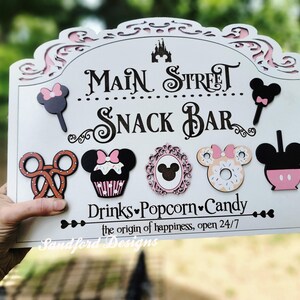 May include: A white wooden sign with the words "Main Street Snack Bar" in black script. The sign features decorative cutouts and images of snacks, including a pretzel, cupcake, and donut. The sign also includes the text "Drinks, Popcorn, Candy".