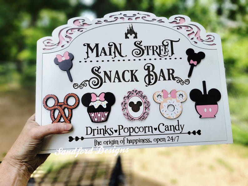 May include: A white wooden sign with the words "Main Street Snack Bar" in black script. The sign features decorative cutouts and images of snacks, including a pretzel, cupcake, and donut. The sign also includes the text "Drinks, Popcorn, Candy".