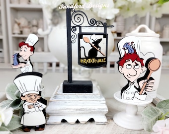 May include: A collection of decorative wooden cutouts featuring characters and a sign from the movie "Ratatouille." The sign reads "La Ratatouille" with a silhouette of a rat holding a spoon. The characters are depicted in chef attire.