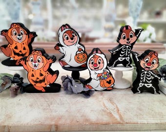 May include: A collection of Halloween-themed Chip and Dale figurines. The figurines are dressed in costumes, including a pumpkin, ghost, and skeleton. The figurines are on small stands and a wooden surface.