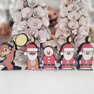 May include: A set of five flat wooden character cutouts featuring a brown chipmunk holding a yellow drum, a reindeer in a Santa hat, a snowman in a Santa suit, and two Santa Claus figures. The text Sandford Designs is printed at the bottom.