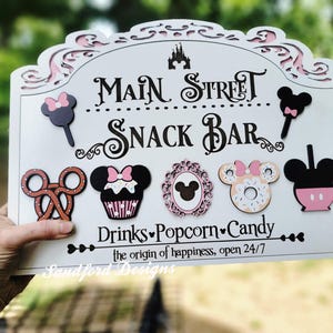 May include: A white wooden sign with the words "Main Street Snack Bar" in black script. The sign features decorative cutouts and images of snacks, including a pretzel, cupcake, and donut. The sign also includes the text "Drinks, Popcorn, Candy".