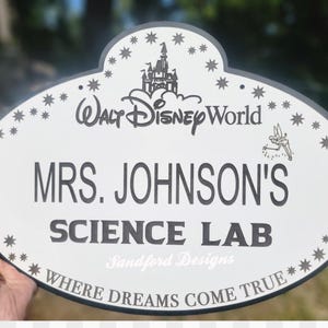 Może przedstawiać: Owalny znak z białym tłem i ciemnoszarą obw&oacute;dką. Znak zawiera tekst "Walt Disney World", "MRS. JOHNSON'S SCIENCE LAB" i "WHERE DREAMS COME TRUE". Zawiera r&oacute;wnież dekoracyjne gwiazdy i grafikę zamku.