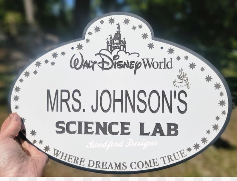 Może przedstawiać: Owalny znak z białym tłem i ciemnoszarą obw&oacute;dką. Znak zawiera tekst "Walt Disney World", "MRS. JOHNSON'S SCIENCE LAB" i "WHERE DREAMS COME TRUE". Zawiera r&oacute;wnież dekoracyjne gwiazdy i grafikę zamku.