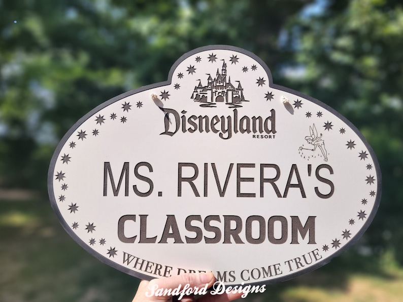 Może przedstawiać: Owalny znak z logo Disneyland Resort, Dzwoneczkiem i gwiazdami. Znak głosi "MS. RIVERA'S CLASSROOM" i "WHERE DREAMS COME TRUE" w ciemnobrązowych literach. Ma białe tło i ciemnoszarą obw&oacute;dkę.