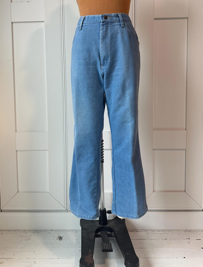 May include: Pale blue denim trousers with a flared leg. The high-waisted jeans have a button and zip fastening. The trousers are displayed on a mannequin against a white backdrop.
