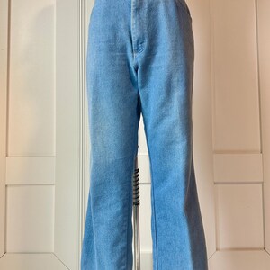 May include: Light blue denim bell-bottom trousers. The trousers have a high waist and a button and zip closure. The legs flare out from the knee to the hem. The trousers are displayed on a mannequin.