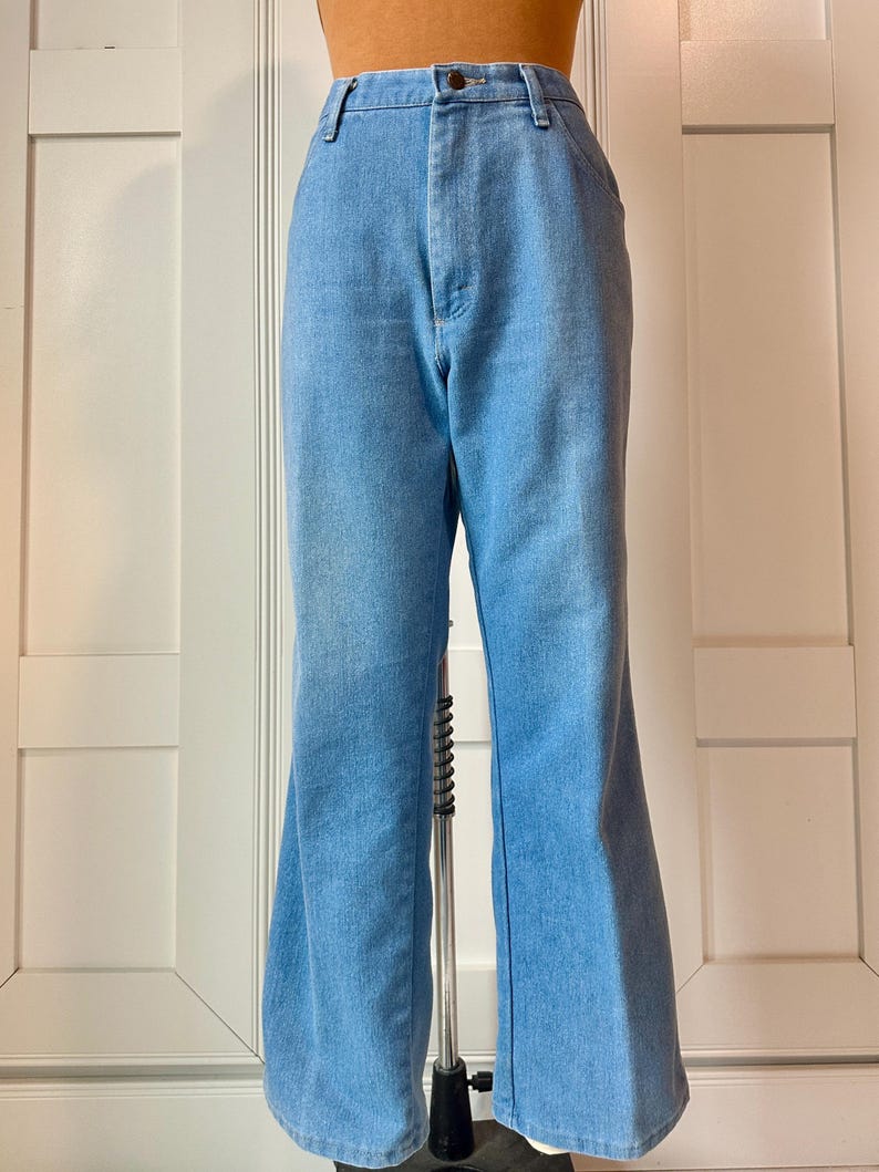May include: Light blue denim bell-bottom trousers. The trousers have a high waist and a button and zip closure. The legs flare out from the knee to the hem. The trousers are displayed on a mannequin.