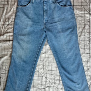 May include: Pale blue denim jeans with a traditional five-pocket design. The jeans feature a straight leg cut and a button and zip fastening. The fabric shows subtle fading, suggesting a vintage style. The jeans are laid flat on a patterned surface.
