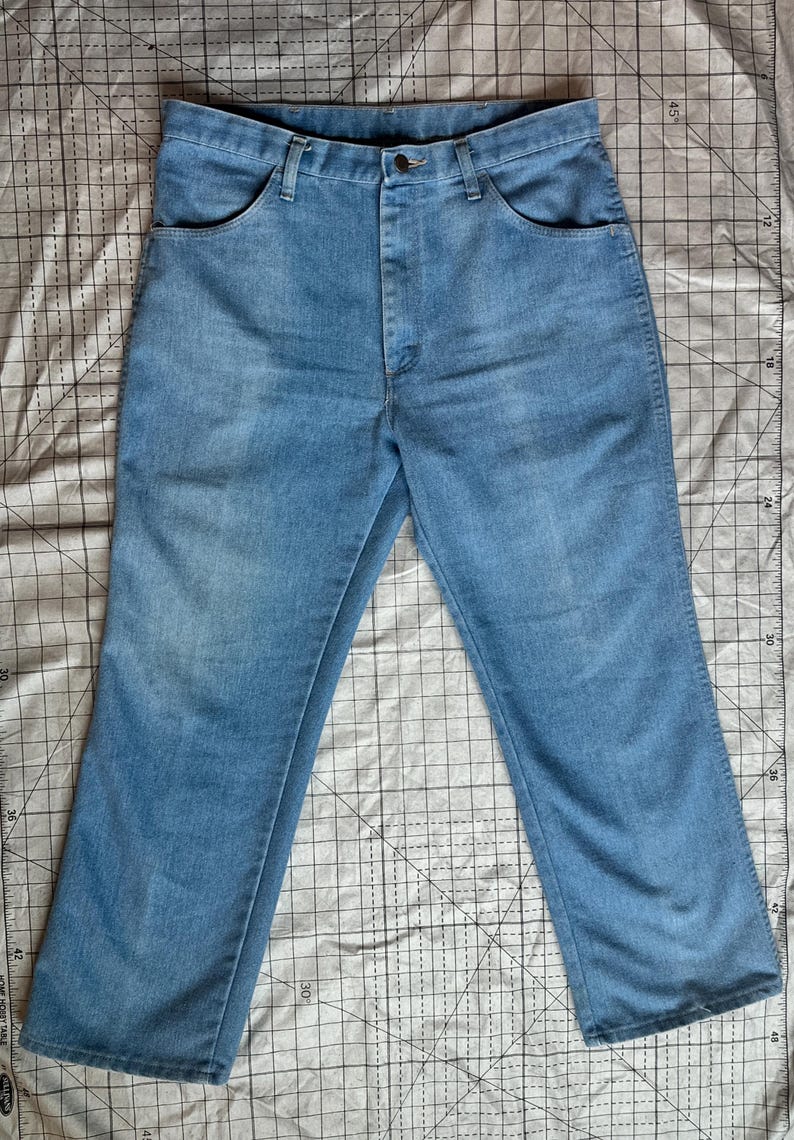 May include: Pale blue denim jeans with a traditional five-pocket design. The jeans feature a straight leg cut and a button and zip fastening. The fabric shows subtle fading, suggesting a vintage style. The jeans are laid flat on a patterned surface.
