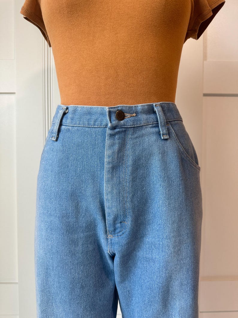May include: Light blue denim jeans with a button closure and belt loops. The jeans are paired with a brown top. The jeans have a classic, straight-leg fit.