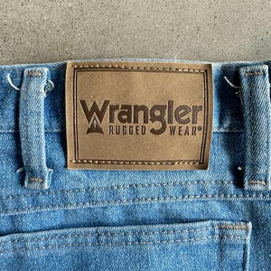 May include: A close-up shot of light blue denim jeans with a brown leather patch. The patch displays the "Wrangler Rugged Wear" logo. The jeans have a classic five-pocket design. The stitching is visible, and the denim has a worn appearance.