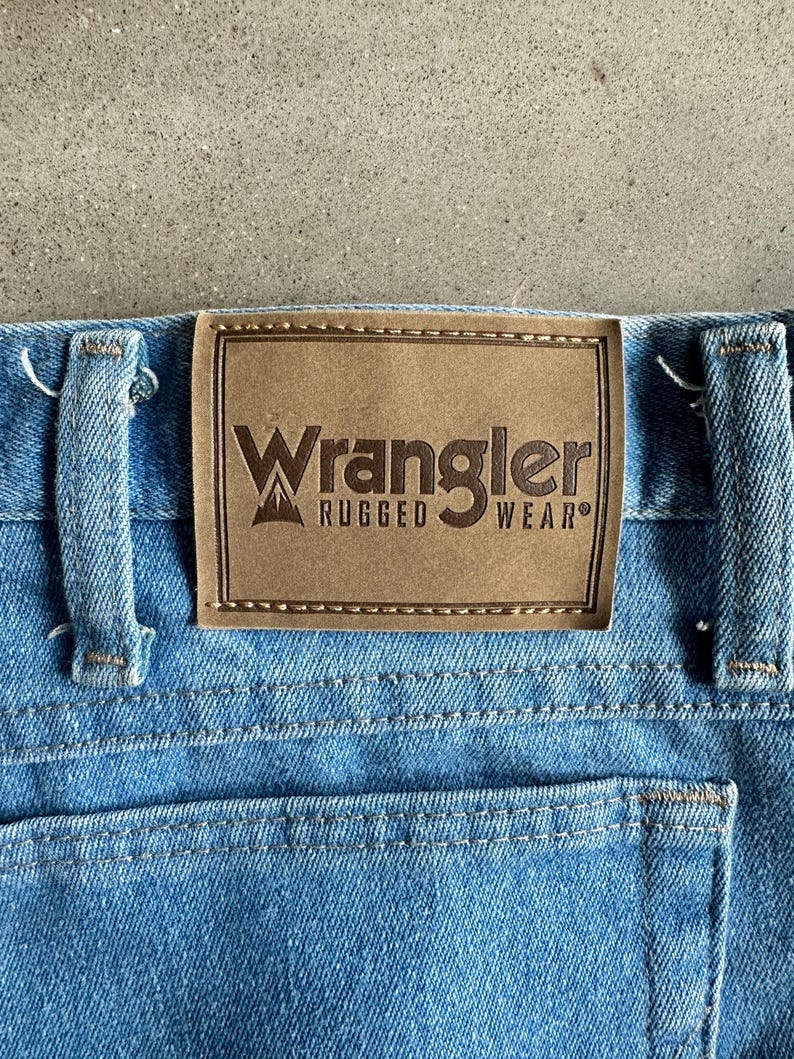 May include: A close-up shot of light blue denim jeans with a brown leather patch. The patch displays the "Wrangler Rugged Wear" logo. The jeans have a classic five-pocket design. The stitching is visible, and the denim has a worn appearance.