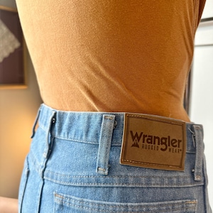 May include: Light blue denim jeans with a brown leather Wrangler label on the back. The jeans have a classic five-pocket design and a straight leg fit. The person is wearing a mustard-coloured top.