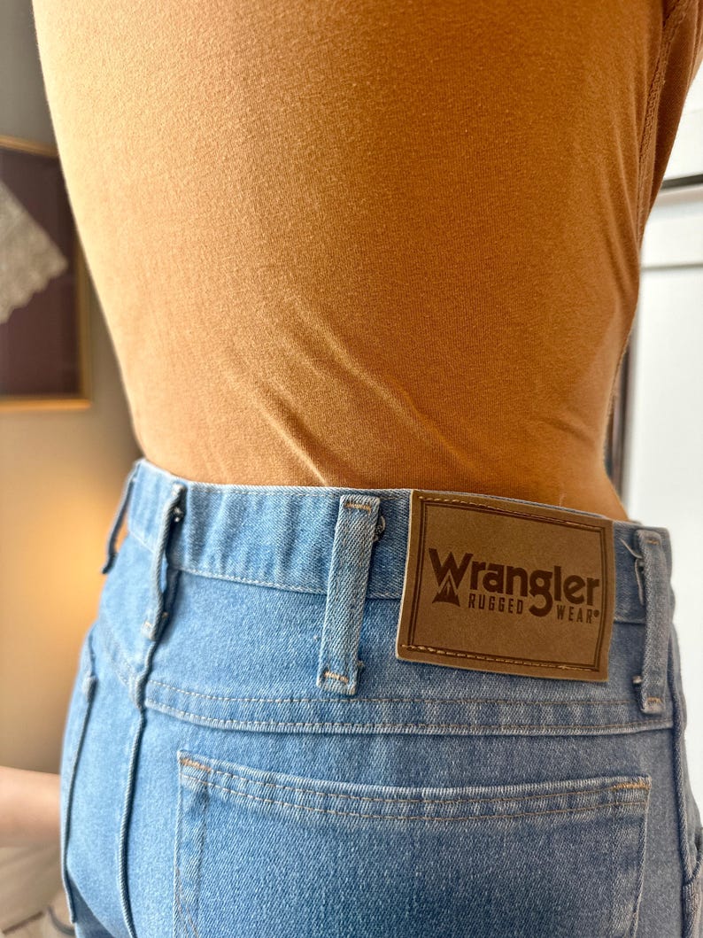 May include: Light blue denim jeans with a brown leather Wrangler label on the back. The jeans have a classic five-pocket design and a straight leg fit. The person is wearing a mustard-coloured top.