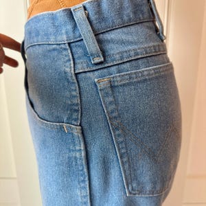 May include: Light blue denim jeans with a classic five-pocket design. The jeans feature a high waist, belt loops, and a back pocket with a stitched V-shaped detail. The denim has a faded wash.