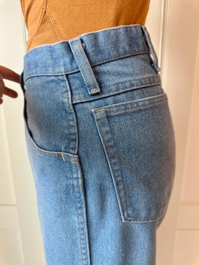 May include: Light blue denim jeans with a classic five-pocket design. The jeans feature a high waist, belt loops, and a back pocket with a stitched V-shaped detail. The denim has a faded wash.