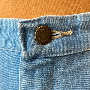 May include: Close-up of light blue denim fabric with a brass button and buttonhole. The button is embossed with the word "Wrangler" in a circular pattern. The denim is stitched with a light brown thread.