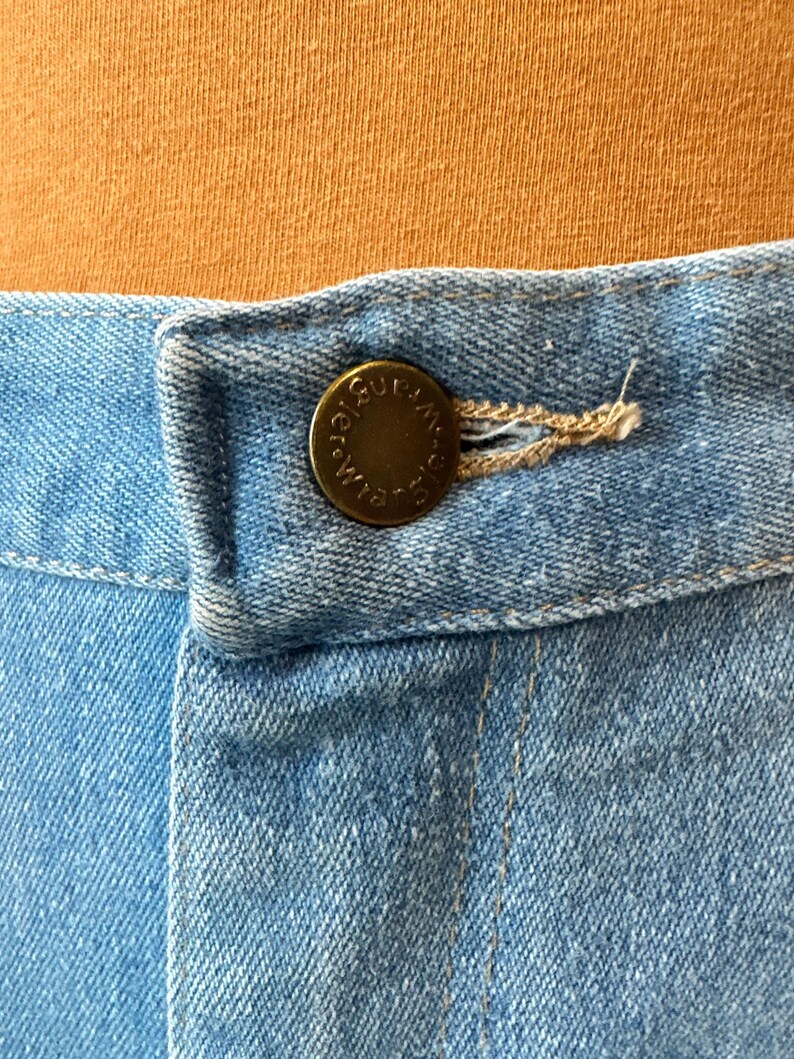 May include: Close-up of light blue denim fabric with a brass button and buttonhole. The button is embossed with the word "Wrangler" in a circular pattern. The denim is stitched with a light brown thread.