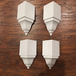 May include: Four white plastic corner moldings with a decorative design. The moldings are arranged in a square pattern on a wooden surface.