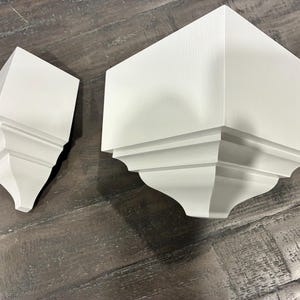 May include: Two white wooden corbels with a smooth matte finish, featuring a square top platform and a curved, stepped architectural profile. These decorative wall brackets are solid in color and display a clean, geometric design with sharp edges.
