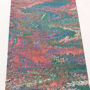 Puede incluir: Un diario hecho a mano con una cubierta veteada. La cubierta presenta un patrón arremolinado de rojo, azul, verde y blanco. Las páginas están encuadernadas con hilo negro. El diario es rectangular y parece nuevo.