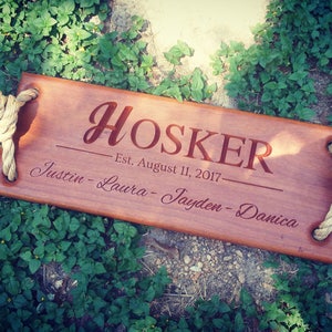 Puede incluir: Un columpio de madera con asas de cuerda, personalizado con el apellido "HOSKER" y el texto "Est. August 11, 2017" junto con los nombres "Justin - Laura - Jayden - Danica".