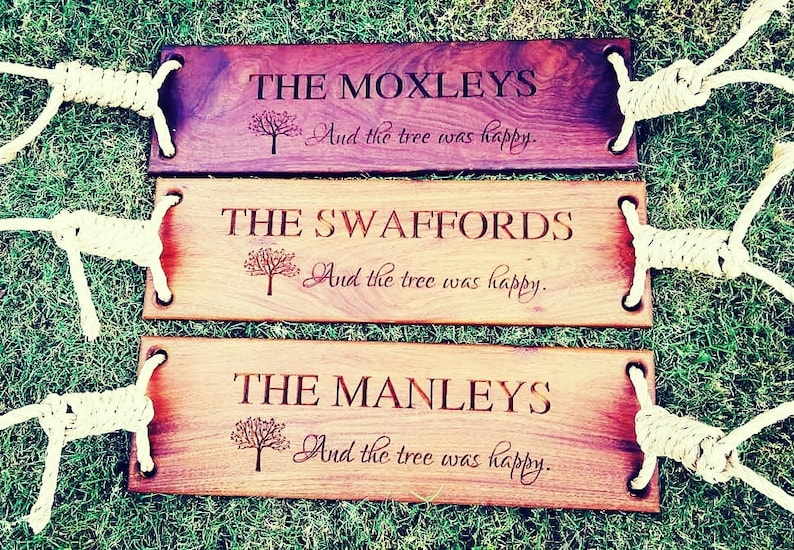 Puede incluir: Tres columpios de madera con asas de cuerda. Cada columpio tiene un nombre personalizado y el texto "And the tree was happy." Los nombres son "The Moxleys", "The Swaffords" y "The Manleys".