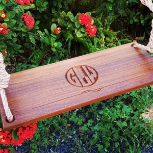 Puede incluir: Un asiento de columpio de madera con una cuerda colgando de &eacute;l. El asiento est&aacute; grabado con el monograma "GLW" en un c&iacute;rculo. El columpio est&aacute; colgado en un jard&iacute;n con follaje verde y flores rojas.