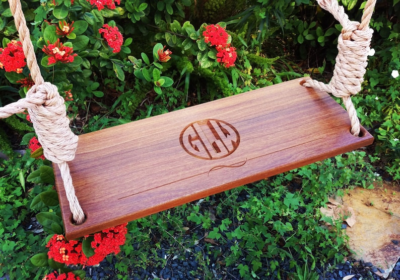 Puede incluir: Un asiento de columpio de madera con una cuerda colgando de &eacute;l. El asiento est&aacute; grabado con el monograma "GLW" en un c&iacute;rculo. El columpio est&aacute; colgado en un jard&iacute;n con follaje verde y flores rojas.