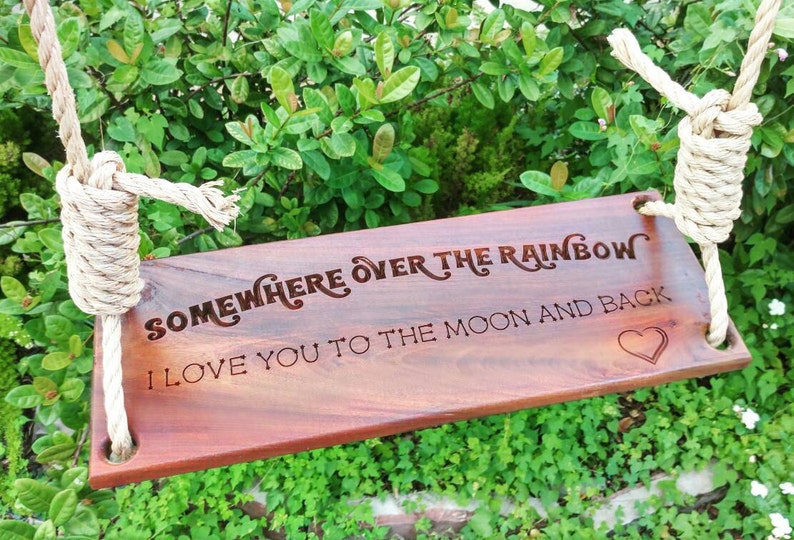 Puede incluir: Un columpio de madera con una cuerda colgando de la parte superior. El columpio tiene el texto "Somewhere over the rainbow I love you to the moon and back" grabado en &eacute;l. Hay una peque&ntilde;a forma de coraz&oacute;n al final del texto.