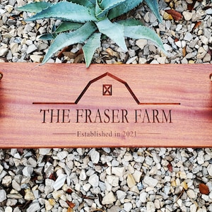 Puede incluir: Un letrero de madera con asas de cuerda, grabado con "THE FRASER FARM - Established in 2021". El letrero presenta una silueta de granero estilizada con una ventana cuadrada.