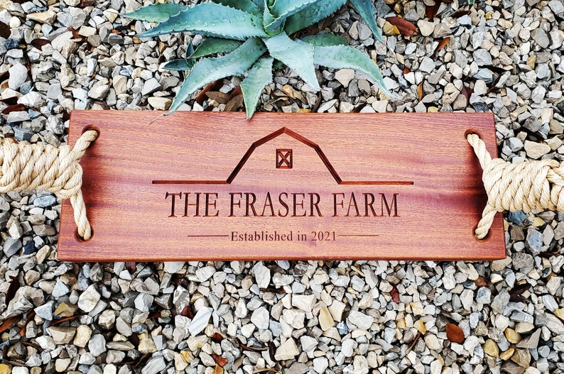 Puede incluir: Un letrero de madera con asas de cuerda, grabado con "THE FRASER FARM - Established in 2021". El letrero presenta una silueta de granero estilizada con una ventana cuadrada.
