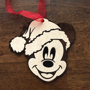 May include: Wooden Christmas ornament featuring a cartoon Mickey Mouse wearing a Santa hat. The ornament is attached to a red ribbon.