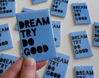 May include: A close-up of a blue square magnet with the text "DREAM TRY DO GOOD" in black lettering. The magnet is being held by a hand.