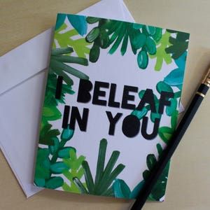 May include: A white card with green foliage printed on the front. The card has black text that reads "I Beleaf in You".