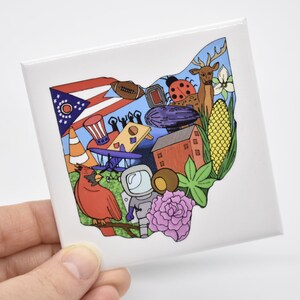 May include: A white ceramic tile with a colorful illustration of the state of Ohio. The illustration includes a cardinal, an airplane, a corn stalk, a ladybug, a deer, a building, a flag, and other symbols.